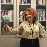 A red haired woman in a colorful shirt holds a globe toy in front of a bookshelf.