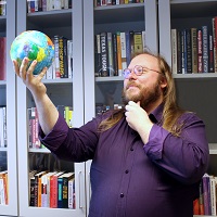 A blond man in a purple shirt holds a globe toy in a theatrical way.