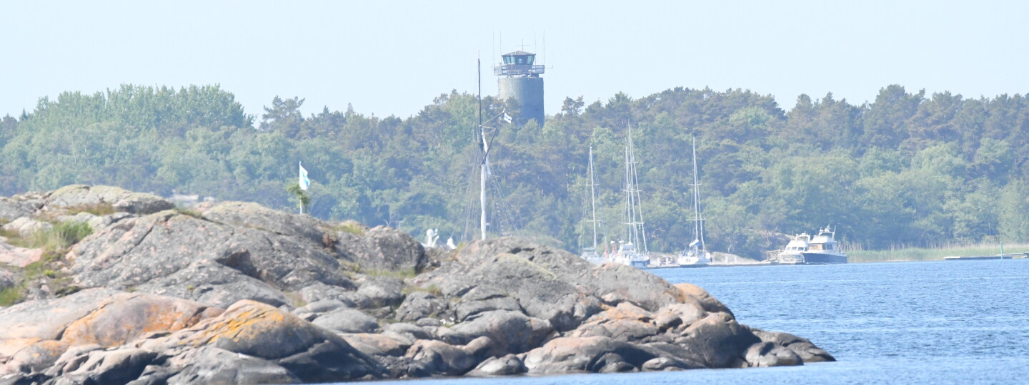 Photo taken from a boat showing blue sea in front, a rocky island behind it and furthest away, the forest like silhouette of the Örö island with a grey watchtower pointing up in the middle. 