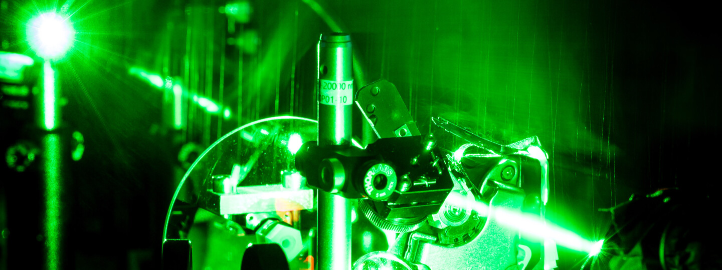 Pimeässä laboratoriossa kuvattu lähikuva vihreästä lasersäteestä läpäisemässä optiikka-laitteistossa näkyviä komponentteja