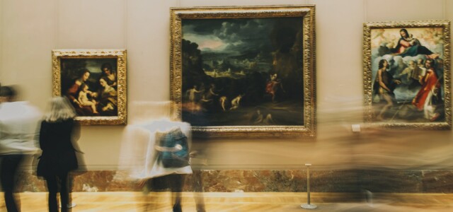 Ihmisiä liikkeessä taidemuseon näyttelytilassa, jonka seinällä on kolme maalausta.