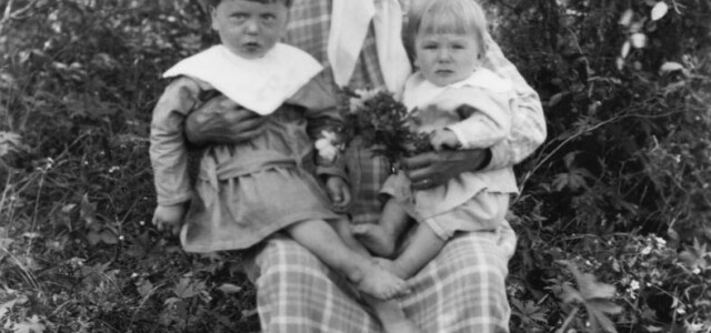 Historiallisessa valokuvassa huivipäinen mummo istuu puutarhassa kaksi pientä lapsenlasta sylissään