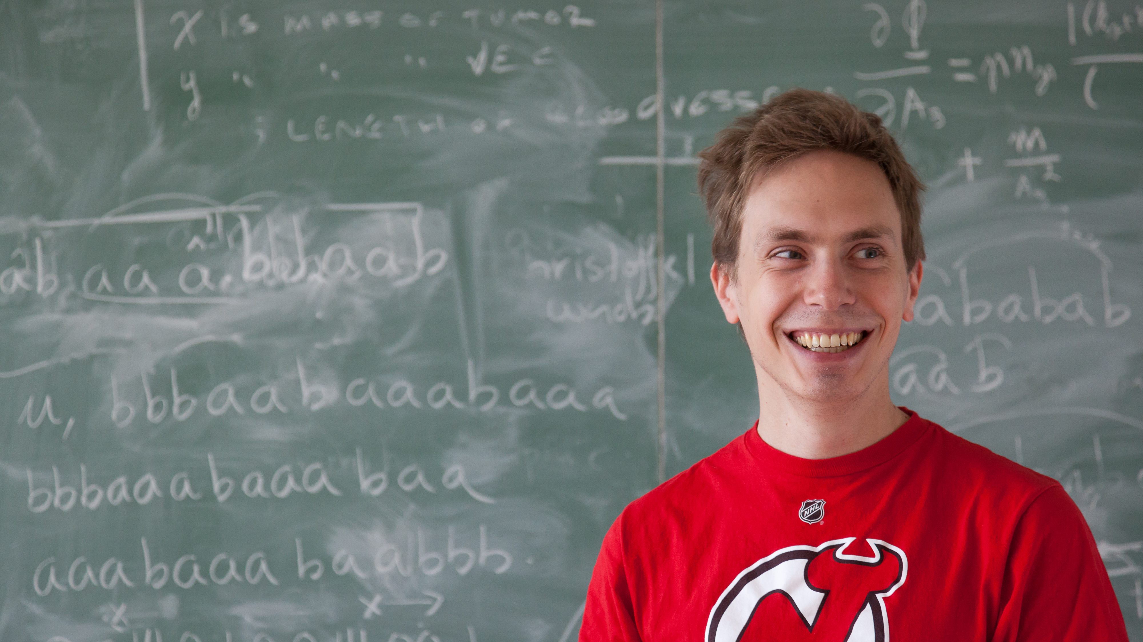 A young man smiling in front of a blackboard filled with mathematical formulas