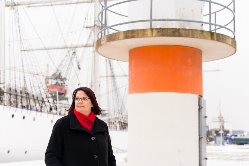 Anne Erkkilä-Välimäki katsoo sivulle ulkona talvella otetussa puolilähikuvassa, taustalla laiva Suomen Joutsen ja pieni majakka.