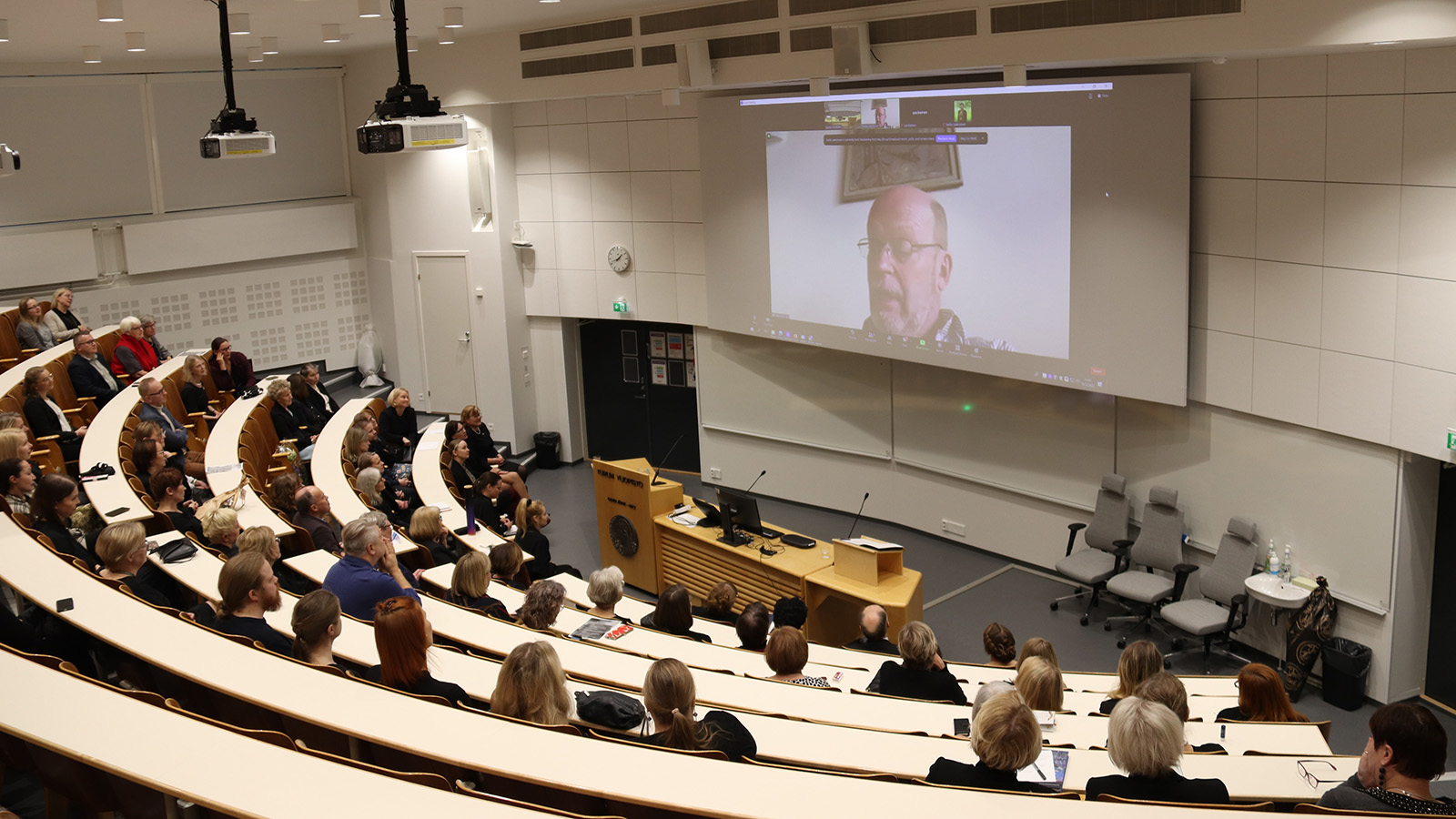 Yleisö kuuntelee Osmo Järvi -salissa, kun dekaani Pekka Hänninen lähettää tervehdyksensä etäyhteydellä. 
