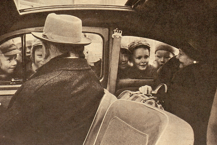 Kaksi ihmistä istuu auton etu- ja takapenkeillä ja auton ulkopuolella ikkunoista katsoo sisään monta lasta.