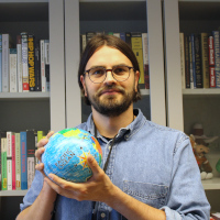 A brunet man in a blue shirt holding a globe toy in front of a book shelf.
