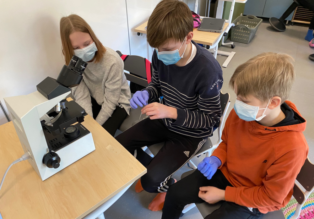 Kolme oppilasta istuu pöydän ääressä ja käyttää mikroskooppia.