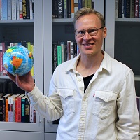 Blond man in a white shirt holds a globe toy in front of a book shelf.