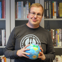 A man holding a globe toy in front of a book shelf and smiling.