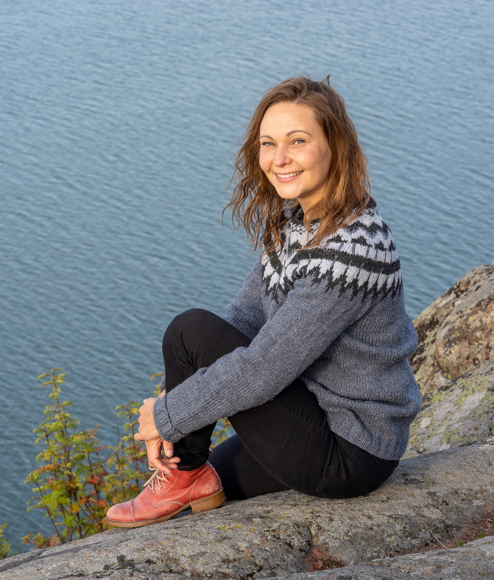 Saija Saarni istuu kalliolla meren äärellä, katsoo kameraan ja hymyilee.