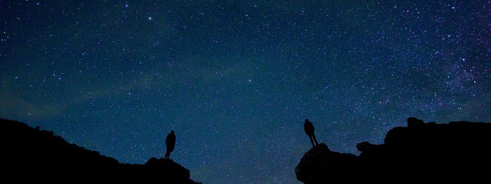 Kaksi ihmistä seisoo pimeässä illassa vuorilla ja katsoo tähtitaivasta.