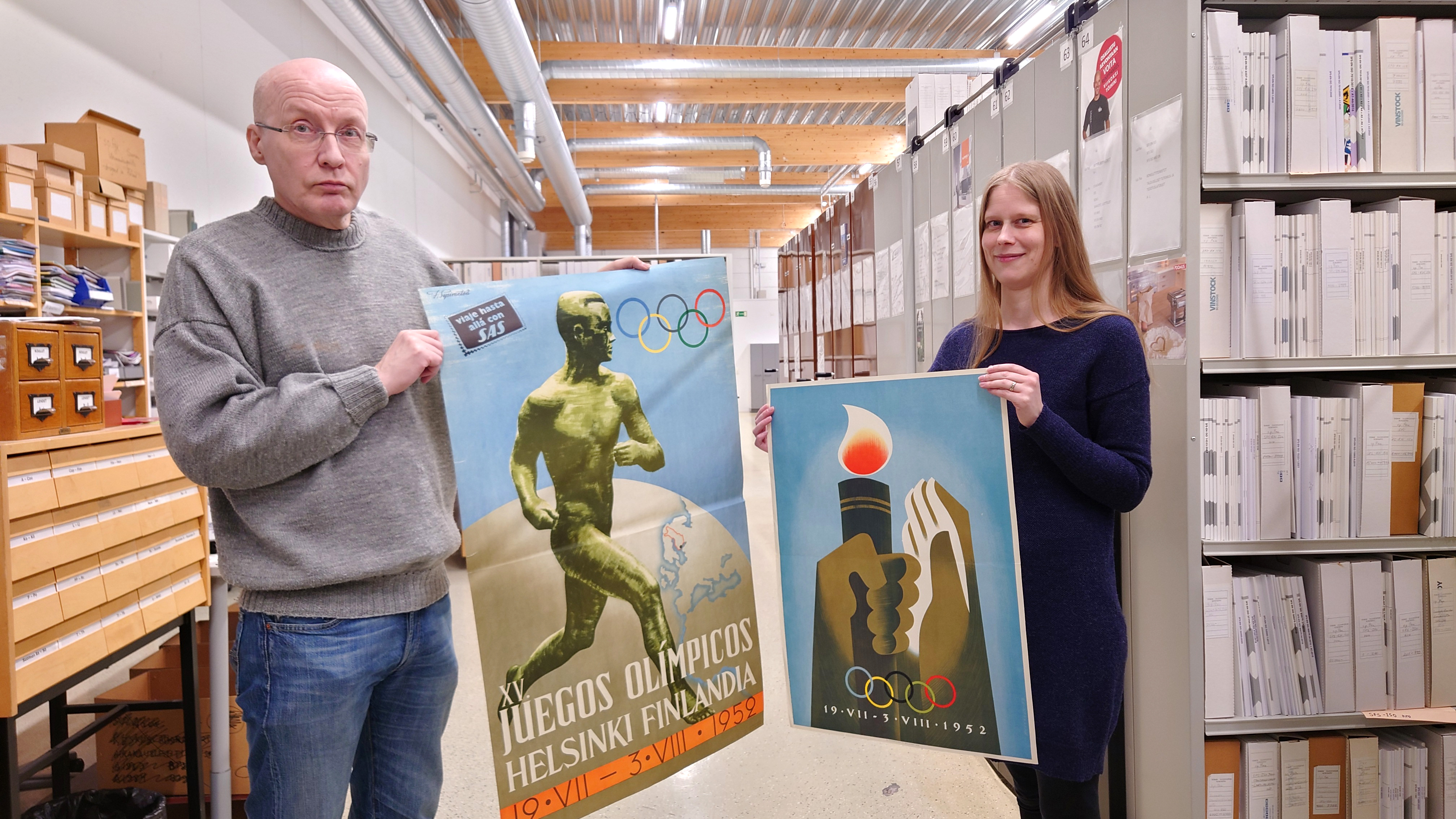 Kaksi kirjastolaista esittelee vuoden 1952 Helsingin olympialaisten mainosjulisteita kirjaston Sanomalehti- ja pienpainatepalveluissa Raisiossa