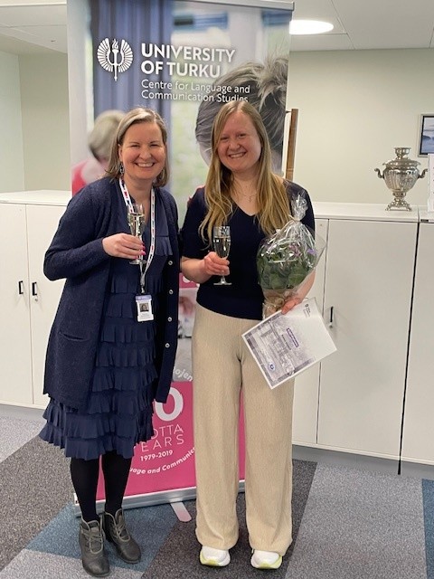 Kaksi henkilöä katsoo kameraan ja hymyilee, käsissään kuohuviinilasit ja toisella myös kukkakimppu ja diplomi.