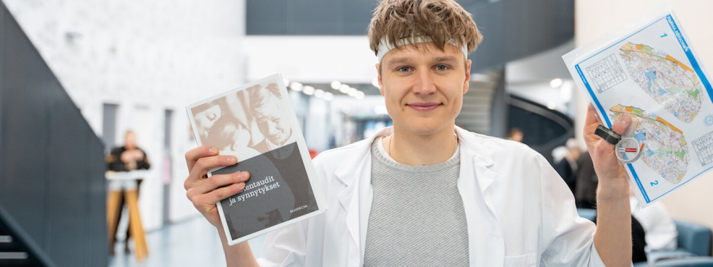 Akseli Ruohola lääkärintakki päällään. Kädessään hänellä on lääketieteellinen opus, kartta ja kompassi