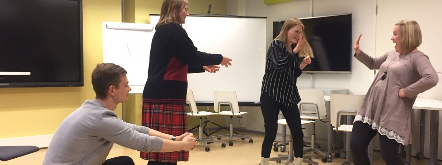 Neljä opiskelijaa harjoittelee vuorovaikutustilanteita iloisin ilmein improvisaatiokurssilla yliopiston opetustilassa.