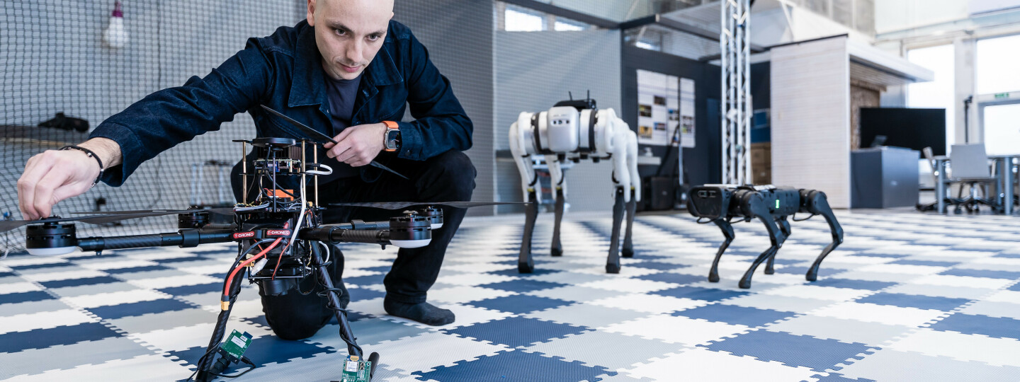 Tutkija kyykistyneenä säätämään dronea, taustalla kaksi robottia