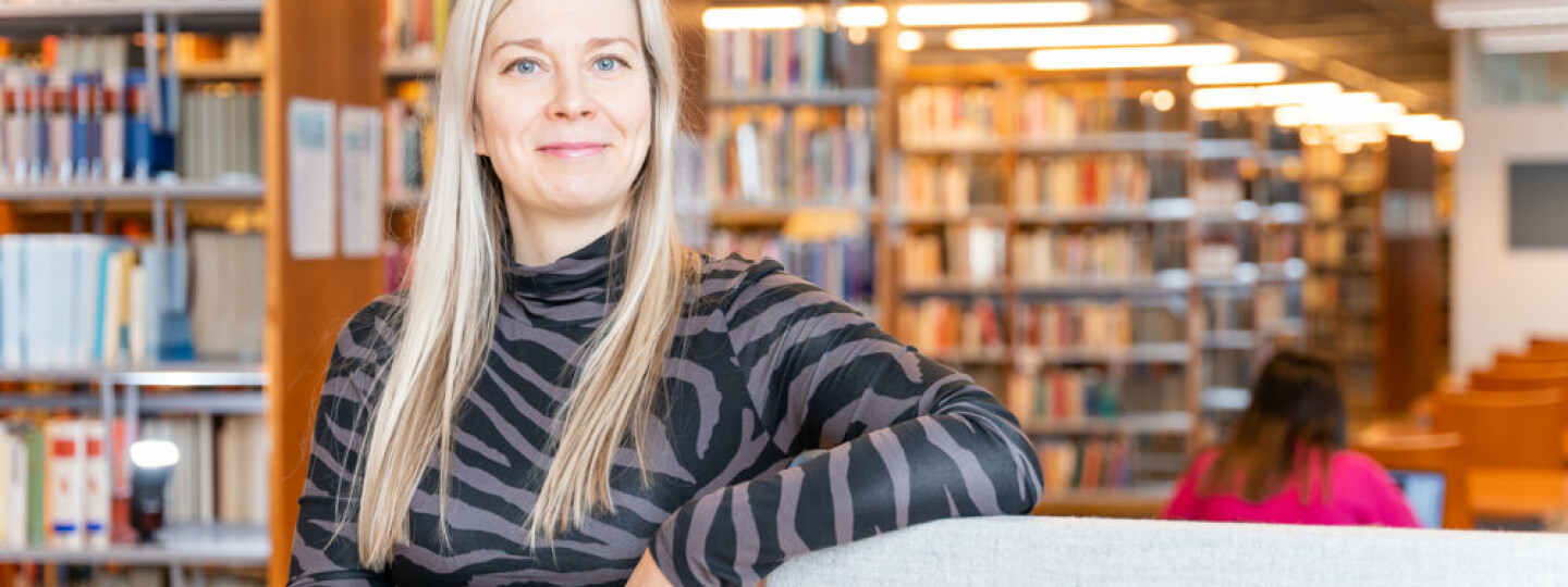 Mirva Heikkilä ketsoo kameraan ja hymyilee puolilähikuvassa, taustalla kirjahyllyjä.