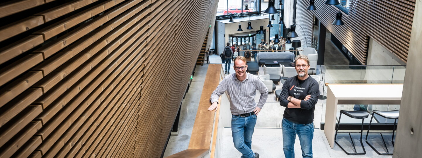 Professors Pasi Virta and Juha-Pekka Salminen are standing on the stairs in the Aurum lobby next to a wooden wall.