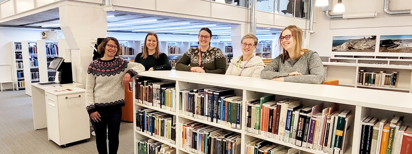 Viisi kirjaston työntekijää seisoo hymyillen matalan kirjahyllyn takana