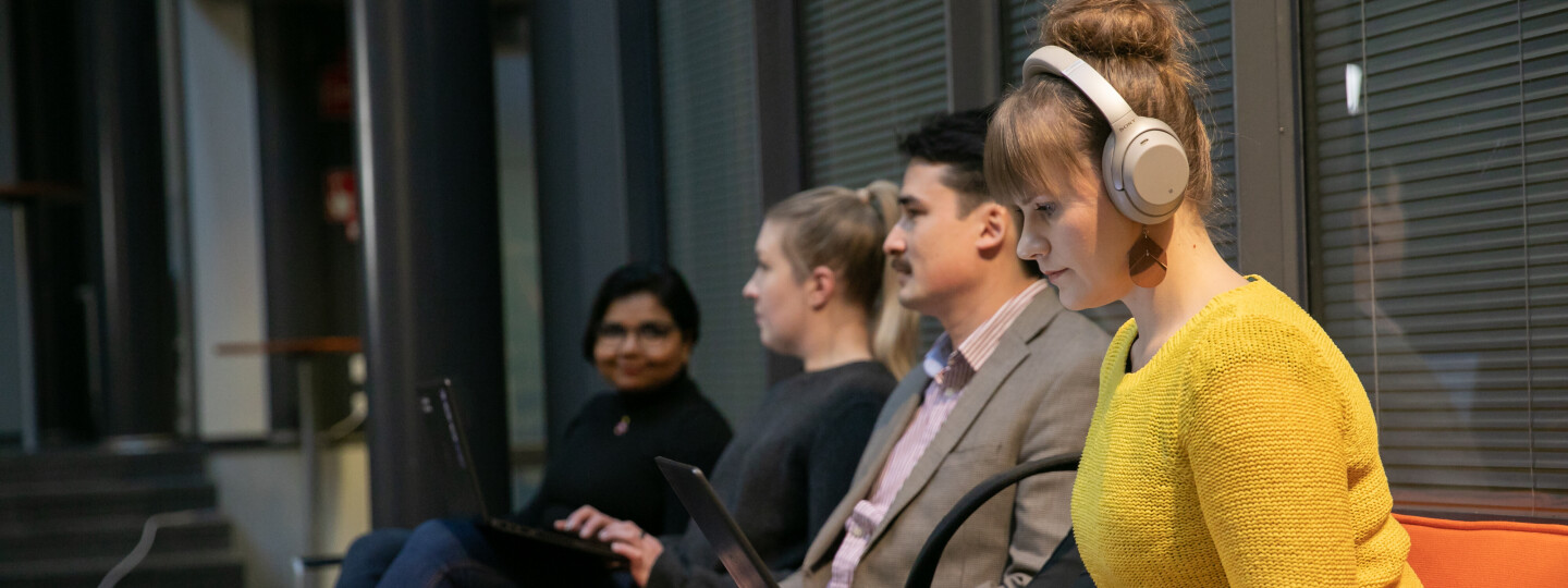 four people sitting in a row, one is wearing headphones and using a laptop