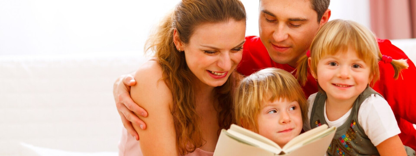 Isä, äiti ja kaksi lasta istuvat sohvalla, äiti lukee lapsille kirjaa