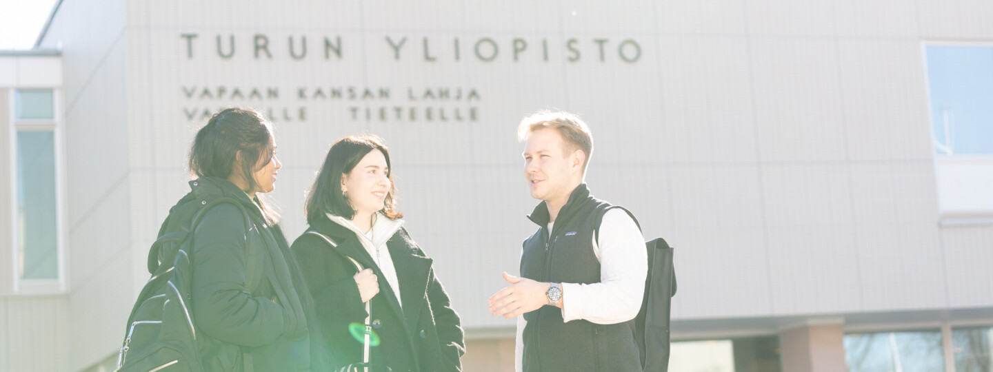 Kolme henkilöä juttelemassa iloisesti Turun yliopiston päärakennuksen edessä. Taustalla yliopiston päärakennuksen seinä, jossa inskriptio "vapaan kansan lahja vapaalle tieteelle".