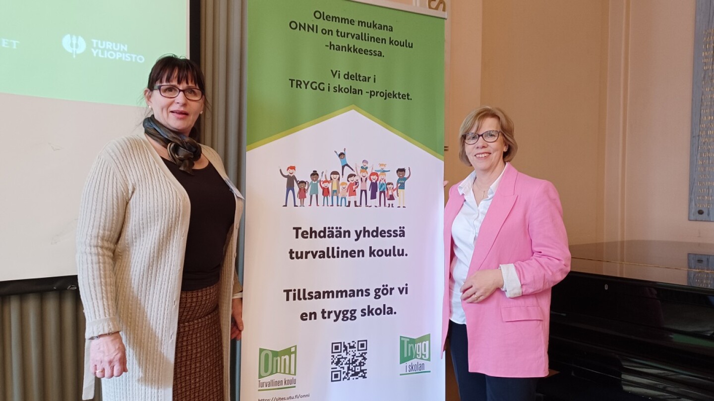 Kaksi naista seisoo kyltin molemmin puolin ja katsoo hymyillen kameraan.