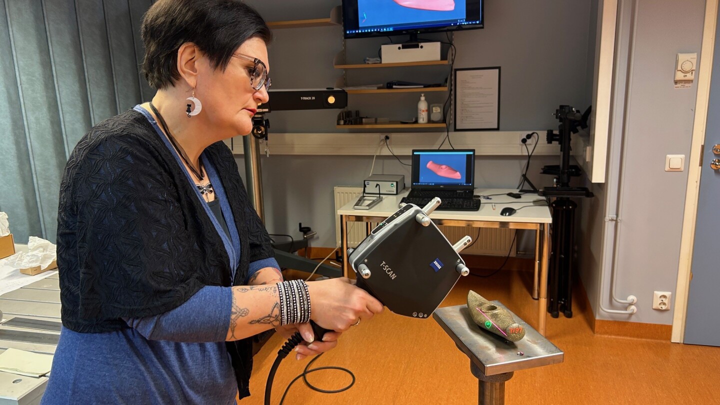 Tutkija pitää digitointilaitetta käsissään ja osoittaa sillä alustan päällä olevaa historiallista vasarakirvestä. Taustalla näkyy läppärin näyttö ja suurempi seinällä oleva näyttö, jonne digitoitu kuva on tullut näkyviin