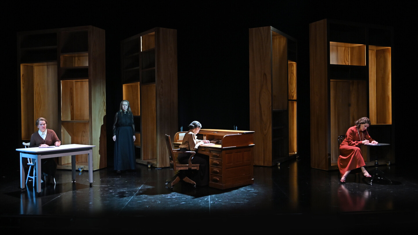 Neljä ihmistä teatterin lavalla, kolme istuu pöytien ääressä kirjoittamassa, yksi seisoo.