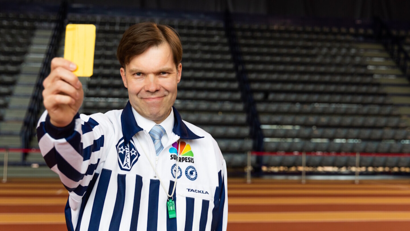 Antti Aine tuomarinvaatteet päällä, näyttää keltaista korttia urheilukentällä.