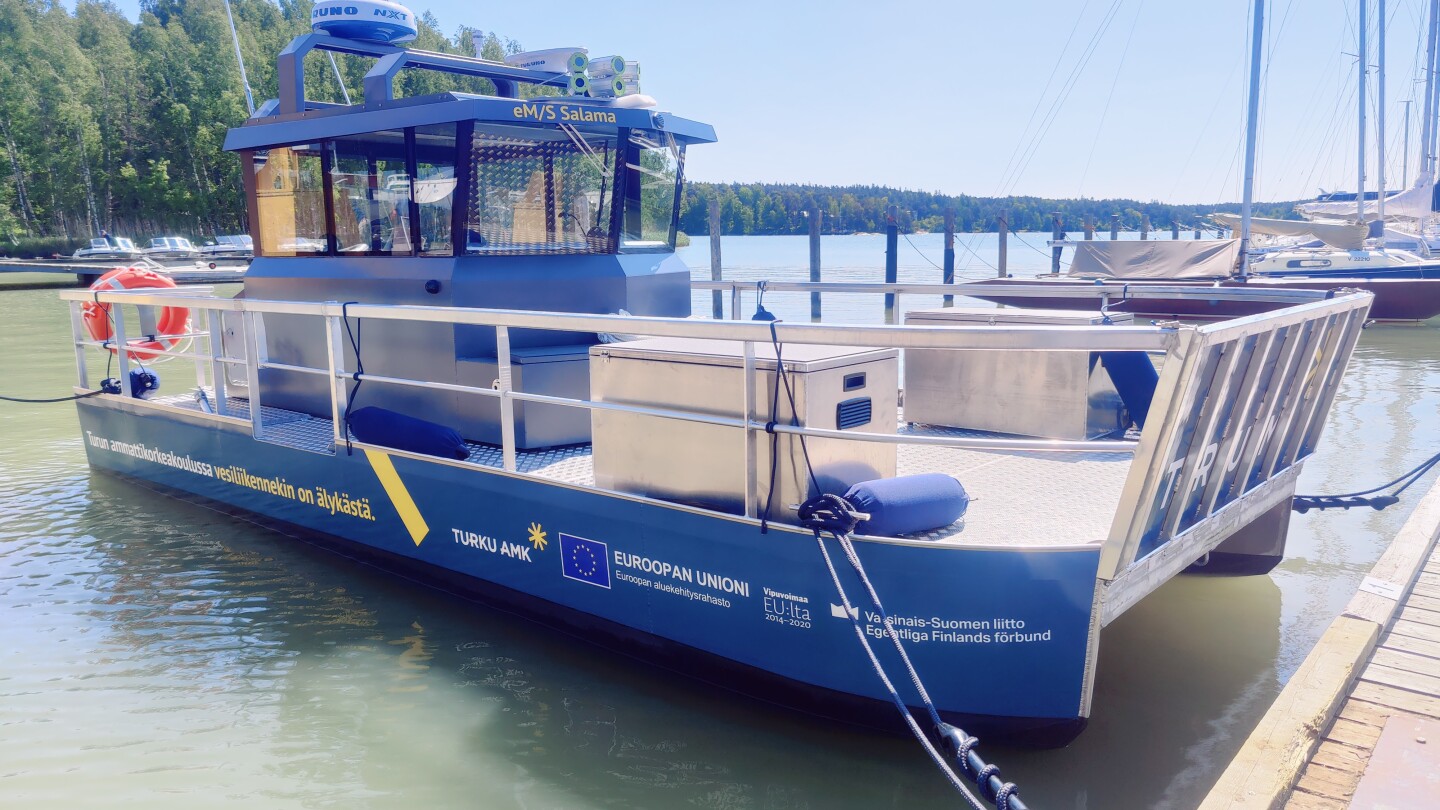 Venesatamassa otettu lähikuva sinisestä testialuksesta, joka on noin 5 metriä pitkä lautta, jota ympäröivät metallikaiteet ja takaosassa sininen ohjaamokoppi.