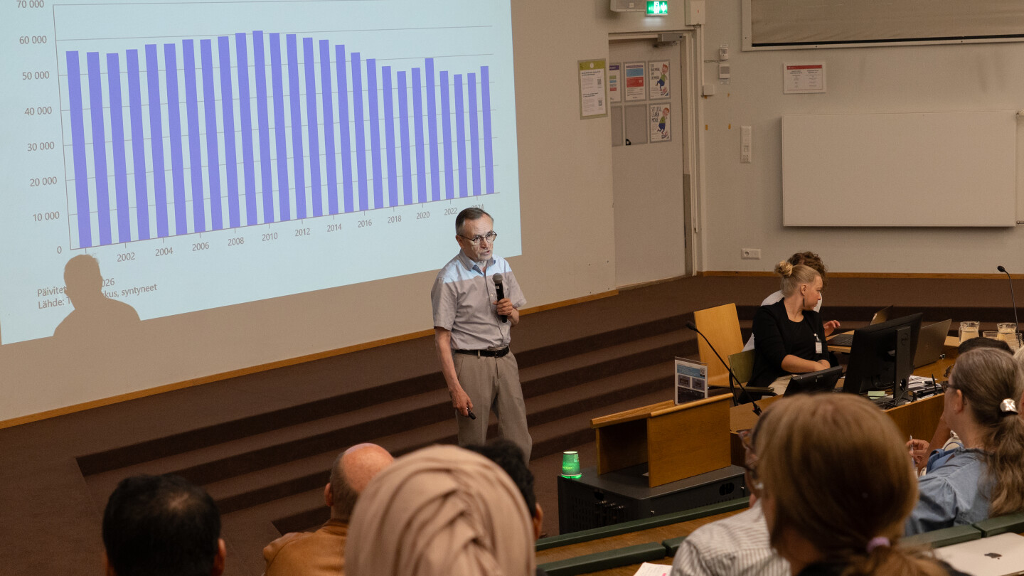 Jorma Toppari puhuu luentosalissa. Valkokankaalla näkyy heijastettyna laskevat syntyvyyspalkit.