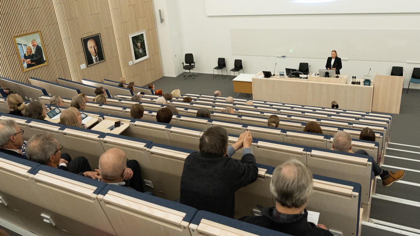 Pirjo Mustonen puhumassa salin edessä, yleisö kuuntelee