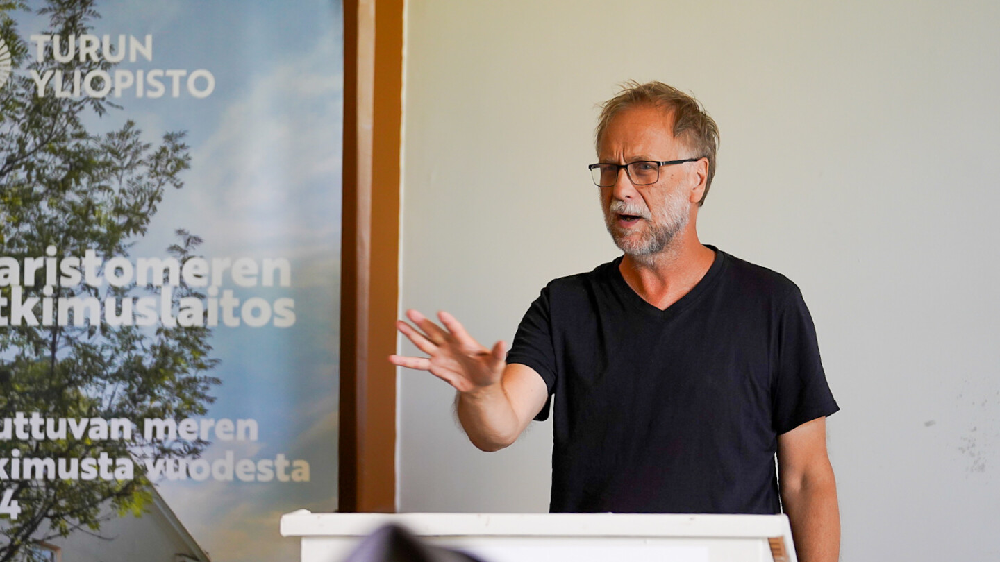 Risto Isomäki seminaarisalin edessä puhumassa yleisölle, käsi nostettuna puhujan eteen painottavana eleenä