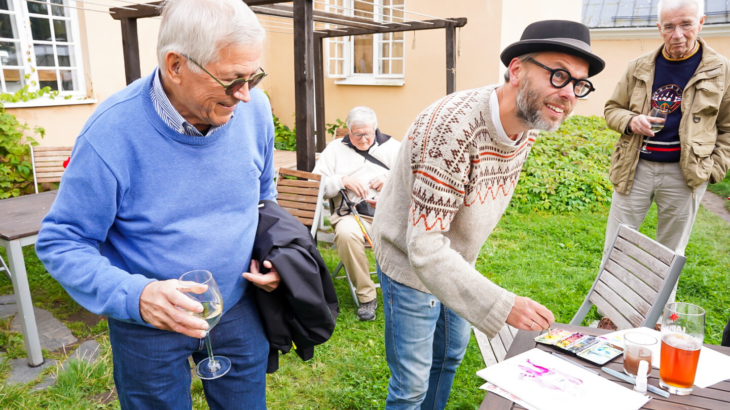 Sarjakuvapiirtäjä Ville Ranta maalaa karikatyyriä Seilin historiallisessa miljöössä kesäisessä pihapiirissä, kumartuneena puutarhapöydän ääreen ja katsoen kohdettaan kameran takana. Ympärillä tapahtuman osallistujia katsomassa Rannan työskentelyä