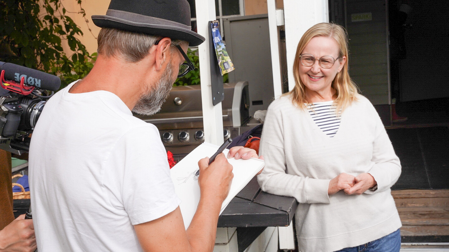 Etualalla selin kuvaajaa kohti sarjakuvapiirtäjä Ville Ranta, hattu päässä ja valkoinen t-paita yllään, pitää toisessa kädessään piirustuslehtiötä ja piirtää karikatyyriä piirtää karikatyyriä kansanedustaja Eeva-Johanna Elorannasta. Eeva-Johanna seisoo Rannan edessä, poseeraa piirtäjälle nojaten kaidetta vasten, ja nauraa iloisesti. Kuvan sivussa näkyy videokameralaitteistoa, kun toimittaja kuvaa tilannetta.