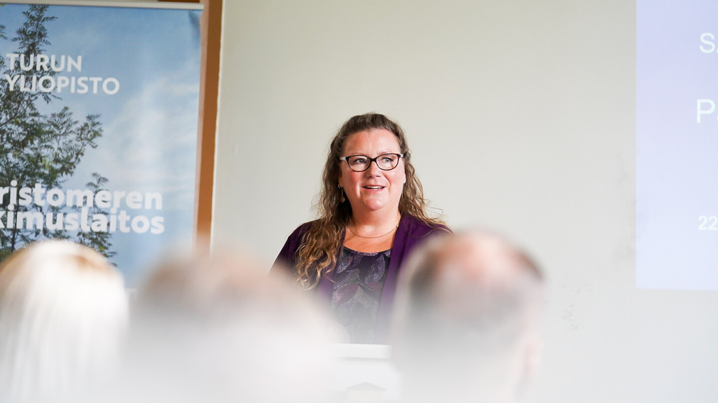 Maija Peltola seminaarisalin edessä pitämässä avauspuheenvuoroa, kuvan alalaidassa etualalla näkyy pari riviä osallistujia takapäin epätarkkana. Puhuja on kauempana salin edessä ja kamera on tarkentanut puhujaan.