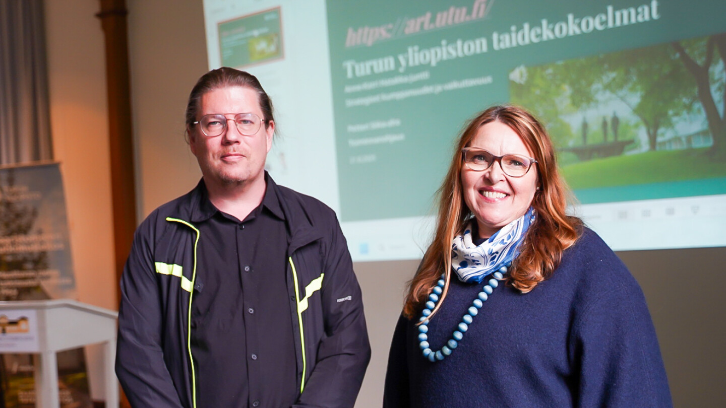 Petteri Siika-aho ja Anne-Katri Hatakka-Juntti vierekkäin seminaarisalissa, molemmat katsovat kameraan ja hymyilevät, taustalla taidegalleriaesityksestä kertova slide heijastettuna salin valkokankaalle