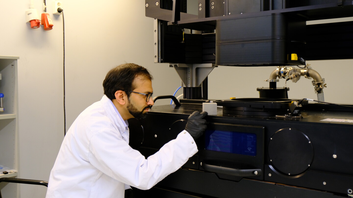 Tutkijalla on yllään valkoinen laboratoriotakki, ja hän on kumartuneena suuren mustan metallintulostuslaitteen äärelle ja tarkastelee sen päällä olevaa esinettä. Laitteen päällä, tutkijan silmänkorkeudella, on kämmenenkokoinen metallikuutio, joka on tulostettu laitteella.