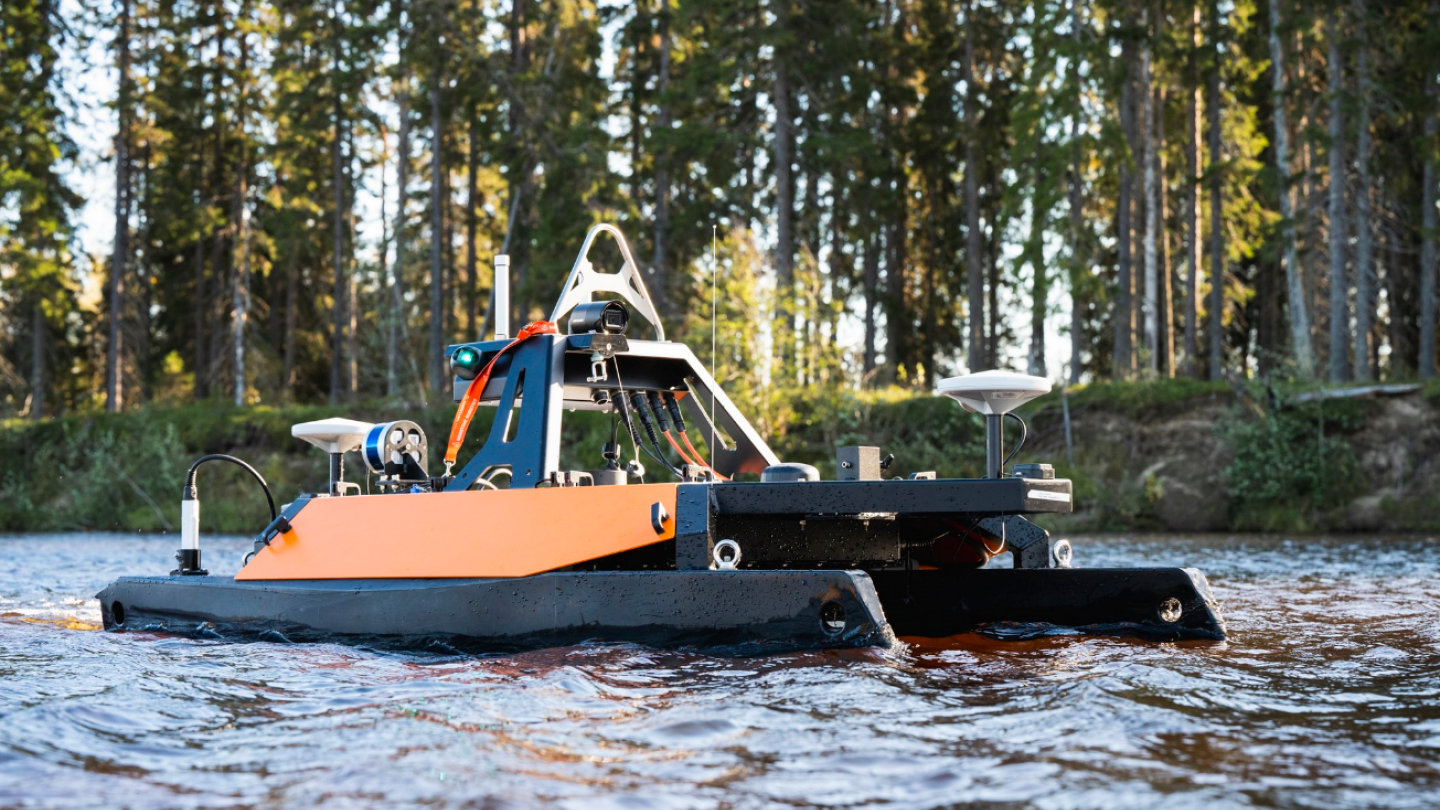 kuvassa etualalla lähikuvassa meressä oleva miehittämätön mittausalus, joka on musta-oranssi etäohjattava pikkukatamaraani, jossa on kiinni mittausvälineita ja instrumentteja. Alus on aivan lähellä rantaa ja takana näkyy mäntymetsää.