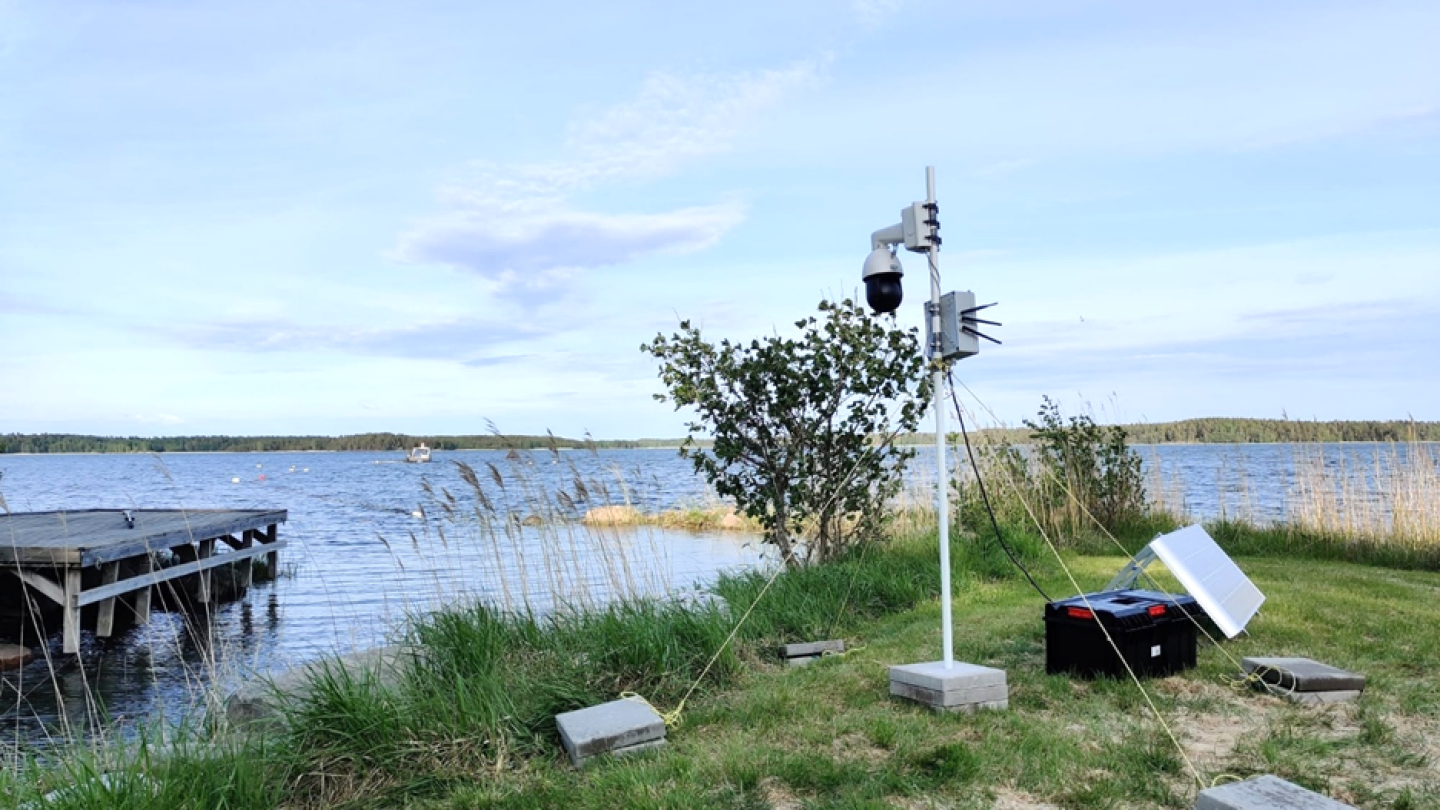 Kesällä merenrannalla otettu kuva rannassa valkoisen pari metriä korkean tolpan päässä olevasta valvontakamerasta, joka kuvaa merellä olevaa kalankasvatusallasta