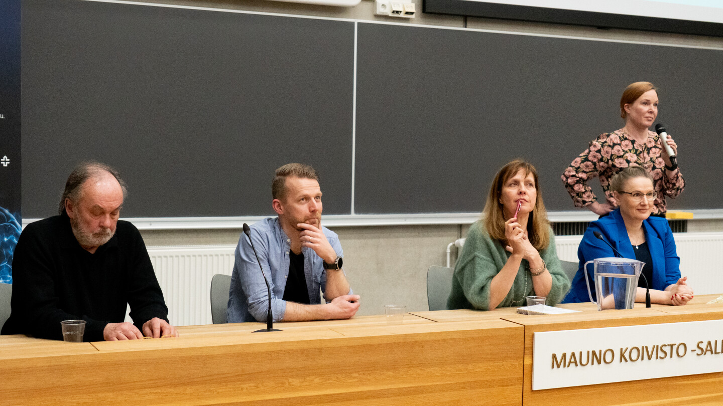 Paneelikeskustelun osallistujat istumassa luentosalin edessä. Takana seisoo juontaja Noora Scheinin