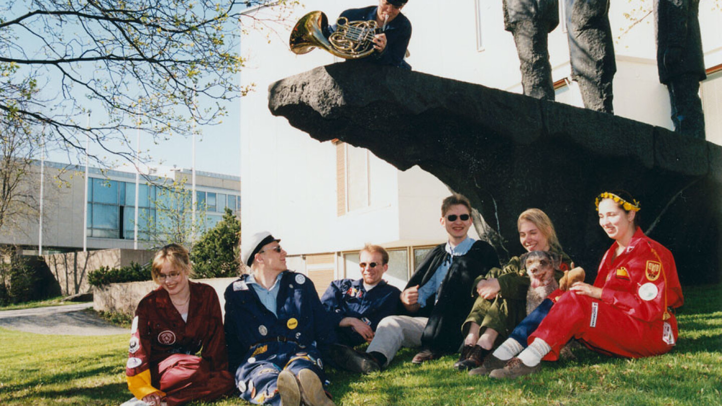 Opiskelijoita istumassa nurmikolla vappuna patsaan alapuolella.