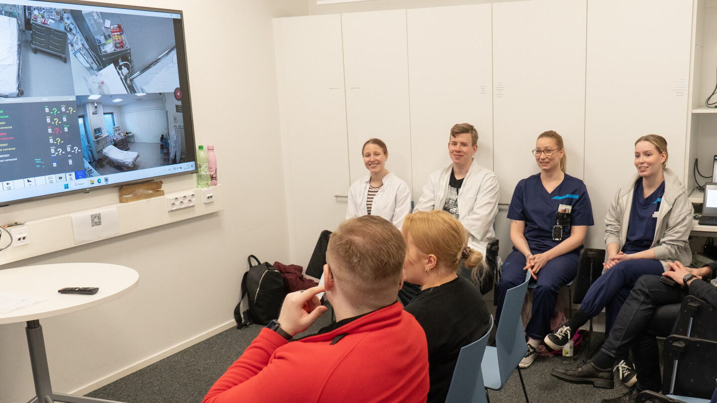 Opiskelijat juttelevat pienessä huoneessa, jonka seinällä on televisio, josta näkyy simulaatiohuone.
