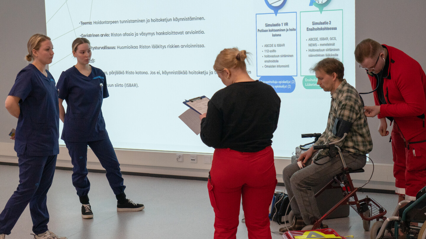 Kaksi sairaanhoitajan vaatteisiin ja kaksi ensihoitajan vaatteisiin pukeutunutta nuorta. Toinen ensihoitaja kuuntelee stetoskoopilla rollaattorissa istuvaa henkilöä.