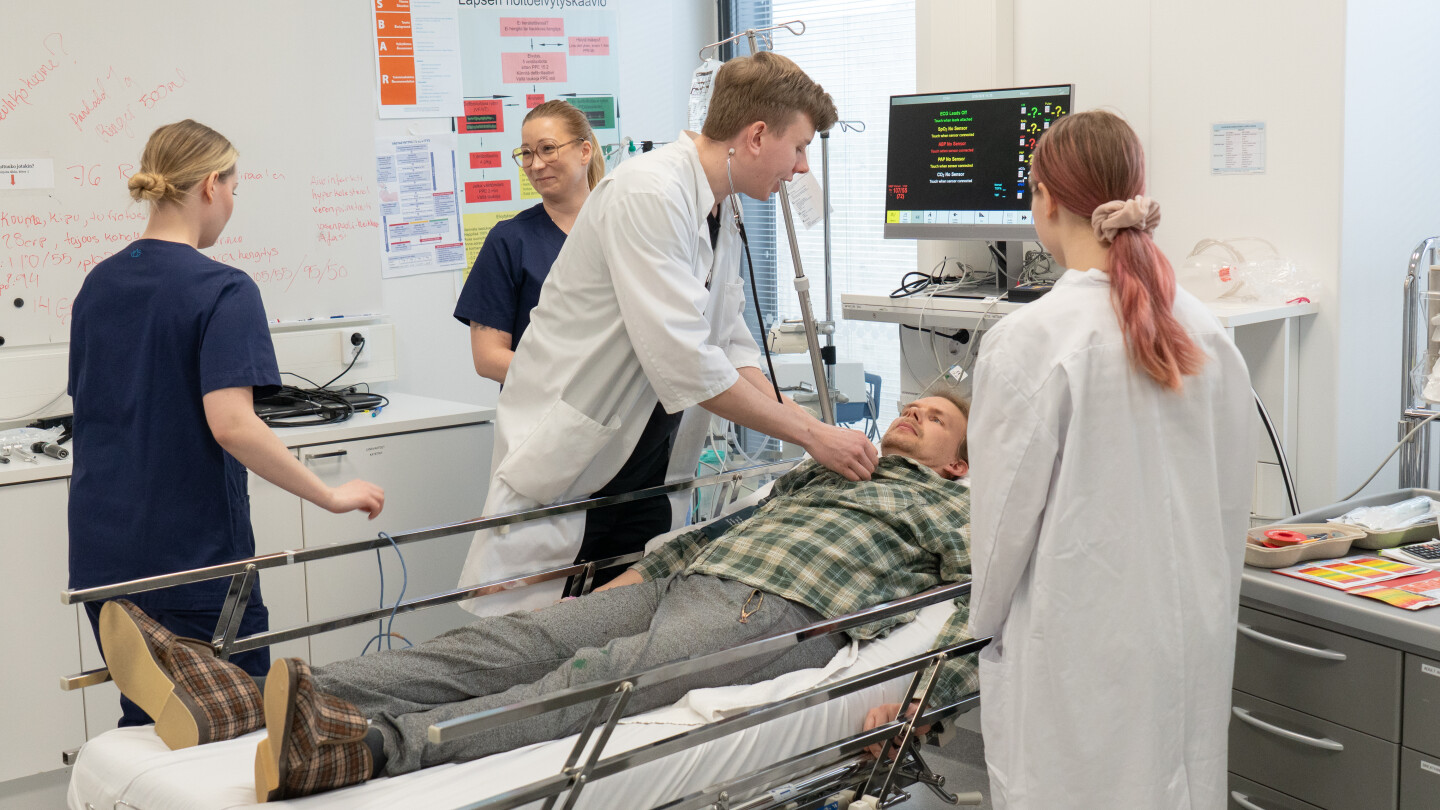 Edustalla kaksi lääkärintakkista nuorta hoitaa sairaalavuoteella olevaan vanhuksen vaatteisiin pukeutunutta henkilöä.Kaksi sairaanhoitajan vaatteisiin pukeutunutta nuorta taustalla.