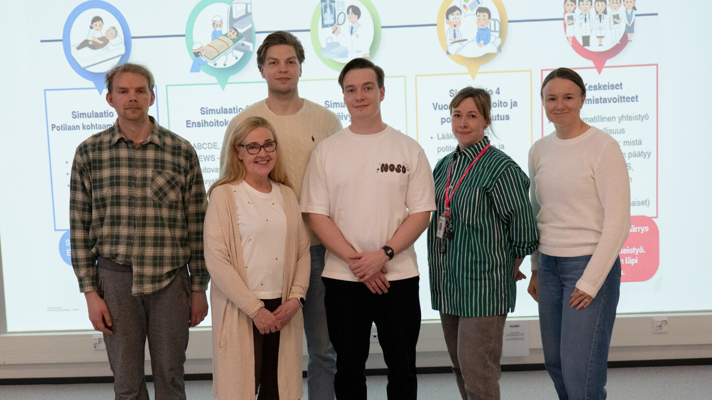 Viisi henkilöä poseeraa kameralle. Taustalla näkyy seinälle hejastettuna simulaation oppimistavoitteita.