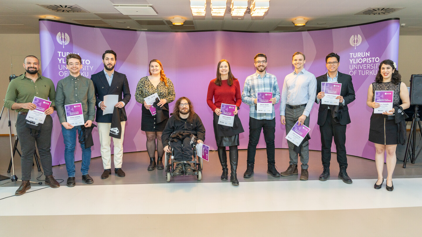 Kymmenen ihmistä seisoo rivissä diplomit käsissään violetin seinän edessä.
