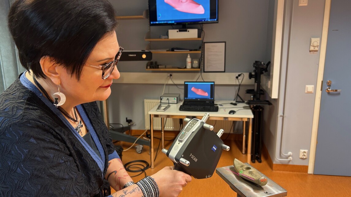 Tutkija pitää digitointilaitetta käsissään ja osoittaa sillä alustan päällä olevaa historiallista vasarakirvestä. Taustalla näkyy läppärin näyttö ja suurempi seinällä oleva näyttö, jonne digitoitu kuva on tullut näkyviin